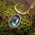 Indlæs billede i Gallery viewer, Et nærbillede af *Vedhæng - Eryn Design - Tranquill 14kt Guld* viser en stor, oval-formet grønlig-blå ædelsten sat i en indviklet sølvring. Vedhænget er lavet af Eryn Design og hænger på en flettet 14 karat guldkæde og hviler på en seng af mos. Ædelstenen reflekterer lyset og giver den et lysende udseende.