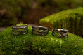 Indlæs billede i Gallery viewer, Udstillet på en seng af frodig grøn mos er tre indviklet designede mørke metalringe med naturinspirerede mønstre, hver prydet med små ædelsten. Disse ringe, kaldet Fingerring - Eryn Design - Moria (Simpel), fremviser en organisk, sammenflettet struktur, der ligner kviste eller grene. De er perfekte eksempler på håndlavede smykker.