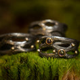 Indlæs billede i Gallery viewer, En trio af mørke, teksturerede ringe fra Eryn Design-kollektionen, med et rustikt, organisk design, hviler oven på en seng af levende grønt mos. To af disse Fingerring - Moria (Simpel) ringe er håndlavede som håndlavede smykker i 925s Sterling Sølv og er prydet med små runde ædelsten sat i gyldne rammer, som tilføjer et elegant touch til den jordiske scene.