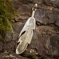 Indlæs billede i Gallery viewer, En Vedhæng Feather 925s Sølv i form af en fjer ligger på groft, barkagtigt træ med noget grønt mos synligt til venstre. Vedhænget har indviklede detaljer, med riller og snit, der efterligner den naturlige tekstur af en fuglefjer vedhæng.