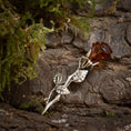 Indlæs billede i Gallery viewer, Broche Rose med Rav 925s Sølv, indviklet formet som en rose, sidder elegant på toppen af struktureret træbark. Dens kronblade er lavet af ægte rav, mens sarte sølvblade strækker sig yndefuldt ud fra stilken. Grønt mos omgiver brochen og forstærker den naturlige skønhed i denne romantiske scene. Et udsøgt stykke perfekt som en romantisk gav.