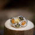 Indlæs billede i Gallery viewer, Sølvring Amber Drops 925s Sølv, med fem forskellige ædelsten arrangeret i et bølget, organisk mønster, vises på en træoverflade. Ædelstenene fremviser forskellige nuancer såsom rav, gul og grøn. Baggrunden er sløret for at holde fokus på det indviklede ringdesign.