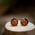 Indlæs billede i Gallery viewer, Et par Ørestikker Amber Key 925s Sølv hviler på en træoverflade. De rige, varme orange-brune sten er indkapslet i en detaljeret sølvramme med små dekorative perler, perfekt fremhævet mod den slørede baggrund.