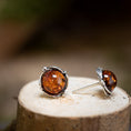 Indlæs billede i Gallery viewer, Et par Ørestikker Amber Key 925s Sølv, med runde rav-sten sat i sterlingsølv, hviler på en naturlig træoverflade. Den ene ørering står oprejst, mens den anden ligger ned og fremhæver det udsøgte håndværk og den rige, rødbrune nuance af ravstenene.