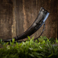 Indlæs billede i Gallery viewer, En elegant Drikkehorn Basic uden holder med en mørk nuance vises elegant på et sort metalstativ, sat mod en træbaggrund. I forgrunden introducerer sløret grønt løv et strejf af naturlig kontrast til den rustikke scene, hvilket fremkalder atmosfæren af traditionel vikingemarkeder.