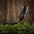 Indlæs billede i Gallery viewer, En smukt udformet Drikkehorn-spiral hviler på et sort metalstativ, sat mod en rustik træbaggrund. Hornet har en skinnende, poleret finish med indviklede spiraludskæringer og er omgivet af frodigt grønt løv i bunden - perfekt til Vikingemarkeder eller som en elegant dekoration.