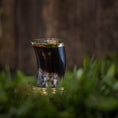 Indlæs billede i Gallery viewer, Et Drikkeglas Horn (100 ml), lavet af poleret dyrehorn og delvist fyldt med væske, er elegant placeret på en træoverflade omgivet af frodigt grønt løv. Baggrunden er sløret, hvilket understreger hornets naturlige, glatte tekstur og reflekterende overflade - et helt unikt drikkeglas.