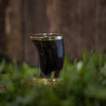 Indlæs billede i Gallery viewer, Et poleret, buet Drikkeglas Horn (100 ml) med en messingkant hviler på en træoverflade omgivet af grønt løv. Baggrunden er sløret, hvilket fremhæver koppens håndværk og de naturlige omgivelser, der ligner et raffineret stykke drikkevare i sin elegance.