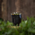 Indlæs billede i Gallery viewer, Et lille, mørkt keramisk Drikkeglas Horn (100 ml) med en skinnende guldkant er placeret udendørs mod en sløret baggrund. Koppens reflekterende overflade viser antydninger af grønt, og forgrunden viser ude af fokus grønne planter, hvilket skaber en effekt, der minder om et smukt drikkeglas.