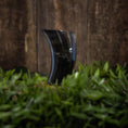 Indlæs billede i Gallery viewer, Drikkeglas Horn Plain (300 ml), et sort, buet glas, der ligner et poleret bøffelhorn, står oprejst på en overflade dækket af grønt løv. Baggrunden består af et struktureret, brunt træpanel.