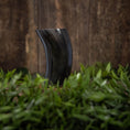 Indlæs billede i Gallery viewer, Drikkeglas Horn Plain (300 ml), et buet drikkehorn lavet af sort bøffelhorn, står oprejst bag en plet med grønt græs. Baggrunden har en rustik træoverflade, der forstærker scenens naturlige og jordagtige atmosfære.