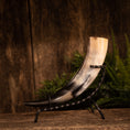 Indlæs billede i Gallery viewer, Drikkehorn Rune med metalholder er et poleret drikkehorn med en gradient, der går fra sort i spidsen til cremefarvet nær åbningen. Den er dekoreret med runegraveringer og fremvises på et elegant sort metalstativ mod en rustik træbaggrund, suppleret med grønt placeret på højre side.
