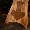 Indlæs billede i Gallery viewer, Et nærbillede af et Drikkehorn Ak m. Thors Hammer Holder, med et detaljeret Thors hammer (Mjölnir) symbol fremhævet med messing søm på nøglepunkter. Træet har en varm tone og fyldig årer, der fremkalder det håndværk, man finder på vikingemarkeder. Baggrunden er blødt sløret med naturlige elementer.