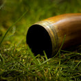 Indlæs billede i Gallery viewer, En smukt udformet Drikkehorn Devine med udsmykkede metaldetaljer på kanten ligger på grønt græs. Dette cylindriske drikkehorn af træ indsnævres mod den ende, der ikke er synlig på billedet. Det indviklede metalværk har mønstrede graveringer, der minder om dem, man fandt på vikingemarkeder.