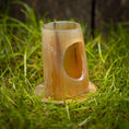 Indlæs billede i Gallery viewer, En cylindrisk, udskåret bambusgenstand med en oval udskæring sidder oprejst på en plet af grønt græs. Bambusen, som hedder "Drikkehorn Devine", fremstår glat med naturlige farvevariationer og er placeret på en lille træbund, der ligner et drikkehorn fra vikingemarkeder.