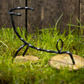 Indlæs billede i Gallery viewer, En Drikkehorn Spiral skulptur i sort metal står på to runde træbaser placeret på græs, med et plankeværk i baggrunden. Figuren, der minder om dekorationer på vikingemarkeder, ser ud til at være i en dynamisk, skæv positur.