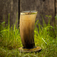 Indlæs billede i Gallery viewer, Et dekorativt Drikkeglas Horn med mønster (300 ml), med en gradient fra mørk til lysebrun og prydet med ætsede geometriske mønstre, vises på en rund træstand midt i grønt græs med et sløret træhegn i baggrunden.