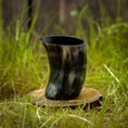Indlæs billede i Gallery viewer, Et Drikkeglas Nature Horn (100 ml), lavet af flerfarvet bøffelhorn, hviler på en rund træbund midt i et græsareal. Baggrunden har et rustikt, sløret udseende, muligvis med træelementer.