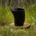 Indlæs billede i Gallery viewer, Drikkeglas Nature Horn (100 ml), et sort, buet drikkeglas lavet af bøffelhorn, står oprejst på en træskranke, omgivet af grønt græs med en sløret naturlig baggrund.