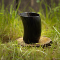 Indlæs billede i Gallery viewer, Et poleret Drikkeglas Nature Horn (100 ml) er placeret oprejst på en træskranke, sat på en plet med grønt græs udendørs. Det unikke glas tilføjer en rustik og jordnær følelse til scenen.