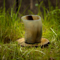 Indlæs billede i Gallery viewer, Et poleret Drikkeglas Nature Horn (100 ml), hviler på en træskranke på en græsklædt overflade. Denne unikke kop har en glat, blank finish med naturlige farvevariationer lige fra mørkebrun til beige. Græs- og træbaggrunden tilføjer en rustik, udendørs følelse.