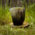 Indlæs billede i Gallery viewer, Et Drikkeglas Nature Horn (100 ml) sidder på en træskranke, omgivet af græs. Koppen har en naturlig gradient fra lys til mørk, hvilket fremhæver dens unikke tekstur og farvevariationer. Billedet er taget udendørs, i rustikke og naturlige omgivelser.