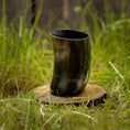 Indlæs billede i Gallery viewer, Et poleret Drikkeglas Nature Horn (100 ml) vises på en træskive midt i højt græs. De naturlige omgivelser understreger hornets rustikke og traditionelle udseende, som har en glat, mørk og blank overflade med lysere striber.