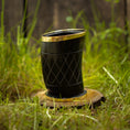 Indlæs billede i Gallery viewer, Et sort Drikkeglas Horn med Mønster (100 ml), indviklet designet med guldkant, hviler på et lille, rundt stykke træ i græsset. Baggrunden viser sløret grønt løv og en utydelig træstruktur, der fremkalder charmen og det unikke ved håndværksfremstillede stykker.