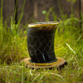 Indlæs billede i Gallery viewer, Et poleret, mørkt Drikkeglas Horn med Mønster (100ml) med en gylden kant sidder oven på en træbase på græs. Koppen har et krydsmønster ætset ind i overfladen og er sat mod en sløret baggrund med naturlige elementer.
