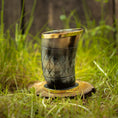 Indlæs billede i Gallery viewer, Et Drikkeglas Horn med Mønster (100ml) med en gylden kant sidder oprejst på en cirkulær træbund i græs. Hornet, der har et diamantmønster og en blank finish, er sat mod en baggrund af sløret grønt og træplanker, hvilket fremhæver dets unikke håndværk.