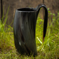 Indlæs billede i Gallery viewer, Et Vikingekrus Nature Horn (600-800 ml), med et slankt, mørkt, buet design med et håndtag, står på en plet græs. Kruset har en poleret, skinnende overflade og en organisk let snoet form. Baggrunden er lidt sløret og ser ud til at være af træ.
