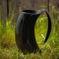 Indlæs billede i Gallery viewer, Et sort Vikingekrus Nature Horn (600-800ml) står på grønt græs med et rustikt plankeværk i baggrunden. Kruset, der efterligner den naturlige form af et dyrehorn, har en slank, blank finish og et stort buet håndtag.