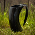 Indlæs billede i Gallery viewer, Et slankt, sort Vikingekrus Nature Horn (600-800ml) med sit unikke buede design står på græs med et plankeværk i baggrunden. Kruset har et utraditionelt håndtag, der passer problemfrit ind i dets moderne æstetik. De naturlige udendørs omgivelser står i smuk kontrast til den moderne stil på denne drikkekrus.