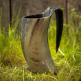 Indlæs billede i Gallery viewer, Et Vikingekrus Nature Horn (600-800ml) står oprejst på den græsklædte grund med et plankeværk i baggrunden. Hornet ligner et traditionelt vikinge-drikkekar og udviser naturlige variationer i farve, der spænder fra mørke til lysebrune nuancer.