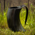 Indlæs billede i Gallery viewer, Et Vikingekrus Nature Horn (600-800ml) med et sort, hornformet design og et buet håndtag sidder på grønt græs, sat mod en sløret træbaggrund. Dette krus efterligner et traditionelt drikkehorn, perfekt til dem, der værdsætter historien og æstetikken ved store drikkekrus.