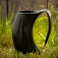 Indlæs billede i Gallery viewer, Et sort Vikingekrus Nature Horn (600-800ml) med et håndtag sidder oprejst på grønt græs, med et træhegn sløret i baggrunden. Hornet, der ligner traditionelle vikingekopper, har en blank finish og buer i en unik form.