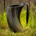 Indlæs billede i Gallery viewer, Et sort Vikingekrus Nature Horn (600-800ml) med en hornlignende form sidder på en græsklædt overflade. Denne unikke håndlavede krus har en blank finish, en bred åbning og et buet håndtag, der ligner et horn. Baggrunden er sløret, hvilket understreger kruset som fokus.