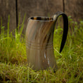 Indlæs billede i Gallery viewer, Et poleret, hornformet Vikingekrus Helleristning Horn (600-800ml) med et håndtag sidder på frodigt grønt græs. Baggrunden er en rustik trævæg, der forstærker scenens naturlige og historiske fornemmelse. Håndlavede hornkrus har subtile linjer og varierende nuancer af brun, tan og sort.
