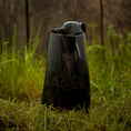 Indlæs billede i Gallery viewer, Et sort keramik Vikingekrus Thors Hammer Horn (600-800ml) med et håndtag og en tud, med et stiliseret Thors hammerdesign ætset på forsiden, er placeret på en plet af grønt græs mod en træbaggrund. Denne vikingekrus udstråler robust charme.