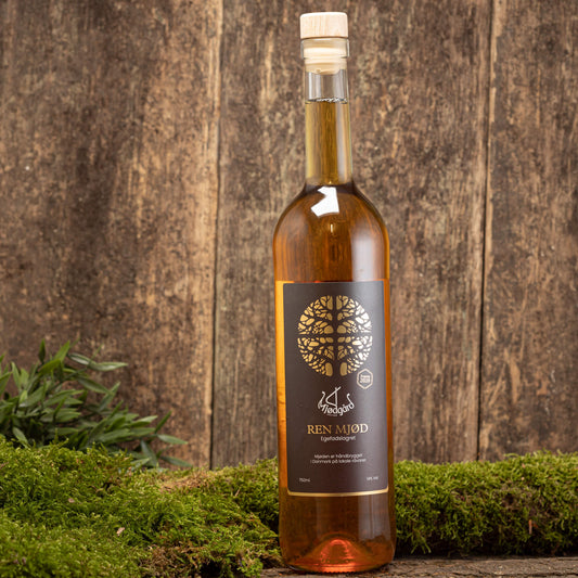 En flaske Mjødgård Ren Mjød (75cl) står på en mosbeklædt overflade. Den mørke etiket har et sølvtrædesign og tekst. Træbaggrunden fremhæver den rustikke æstetik, mens den rige ravvæske indeni minder om fin dessertvin.