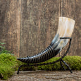 Indlæs billede i Gallery viewer, Drikkehorn-hjorten, der har et poleret horn med en gradient fra sort i bunden til beige i spidsen, er elegant monteret på et sort smedejernsstativ og hviler på en mosbeklædt træoverflade mod en rustik træbaggrund - ideel til vikingemarkeder.