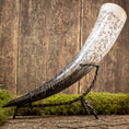 Indlæs billede i Gallery viewer, En dekorativ Drikkehorn Pikto med indviklede udskæringer hviler på et sort metalstativ. Hornet har forskellige runesymboler og design ætset ind i dets overflade. Placeret på en rustik træbaggrund med pletter af grønt mos, fremkalder den atmosfæren af vikingemarkeder.