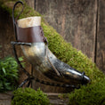 Indlæs billede i Gallery viewer, Et raffineret Drikkehorn Raw med Brun Læderstrop, elegant præsenteret på et snoet metalstativ. Baggrunden viser en rustik træoverflade prydet med grønt mos og løv, der forstærker den naturlige æstetik og fremkalder charmen ved vikingemarker.