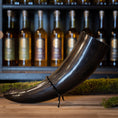 Indlæs billede i Gallery viewer, En poleret sort Drikkehorn Giant 1,5 liter hviler på et stativ foran en hylde fyldt med flasker med forskellige alkoholer, hver prydet med et guld cirkulært logo. Moss dekorerer træoverfladen omkring drikkehornet og tilføjer et rustikt touch til displayet, der minder om vikingemarkeder.