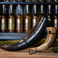 Indlæs billede i Gallery viewer, To udsøgt udformede drikkehorn, inklusive Drikkehorn Giant 1,5 liter, i forskellige størrelser og farver vises på et træbord. Hver bøffelhorn hviler på et metalstativ, og en række flasker er synlige i baggrunden på hylder. Rammerne er rustikke og elegante, der minder om traditionel vikingemarkeder.