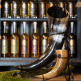 Indlæs billede i Gallery viewer, En udstilling af to drikkehorn, det ene med det imponerende Drikkehorn Giant 1,5 liter i sort bøffelhorn og det andet mindre og brunt, sat på et stativ med en mosbeklædt bund. Baggrunden byder på hylder fyldt med forskellige drikkevarer på flaske, der minder om vikingemarkeder.