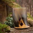 Indlæs billede i Gallery viewer, Et poleret Drikkeglas Horn Plain (500 ml) med et buet design og træbund hviler på en mosbeklædt træstamme. Baggrunden viser en rustik træoverflade, delvist prydet med grønt mos og løv.
