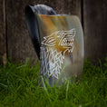 Indlæs billede i Gallery viewer, Et poleret Vikingekrus ulvehorn (600-800 ml), med et stiliseret ulvehoveddesign, der er indviklet udskåret på siden, står oprejst på grønt græs. Baggrunden er lavet af træplanker. Dette unikt designede horn viser en blanding af mørke og lyse nuancer.