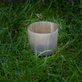 Indlæs billede i Gallery viewer, En opretstående Lysestage til fyrfadslys, cylindrisk og gennemskinnelig, placeret på en plet af grønt græs. Lignet til en delikat messingholder virker den tom og har en glat overflade med en let glans.