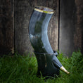 Indlæs billede i Gallery viewer, Drikkehorn Ordained, et poleret bøffelhorn drikkehorn prydet med metalkant og spids, hviler elegant i sit matchende sorte stativ på græs, mod en rustik trækulisse, der minder om vikingemarkeder.